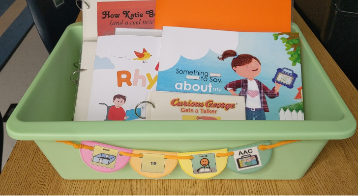Plastic bin with books featuring AAC. The titles that are partially visible are “Curious George Gets a Talker”, Something to Say about my Communication Device”, “AAC Rhyme Time” and, “How Katie Got a Voice (and a cool new name)”. 
