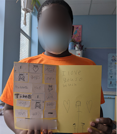 student with a blurred-face in an orange shirt shows off the inside of a yellow paper card he made