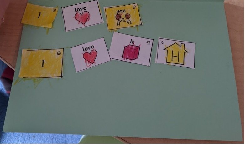 a green paper card with cut-out SymbolStix icons forming a 2-line message. The top line reads, “I love you”. The second line reads, “I love it home (home button)”.