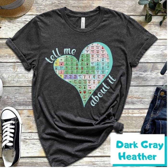 a dark heather gray shirt with a knot on the right side is lying on a wood background. There is a large lopsided heart in the center filled in with a TouchChat core board. On the top left of the heart are the words, “tell me”. Under and to the right of the heart are the words, “about it”.