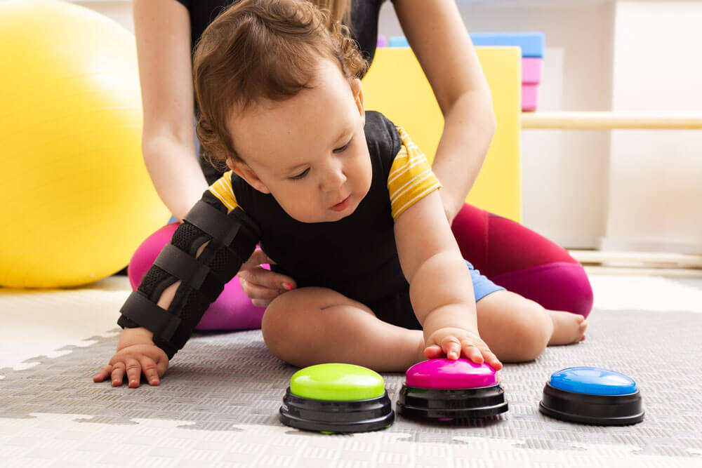 a toddler sitting in front of a therapist on the floor reach toward one of three single message voice output aides in front of them