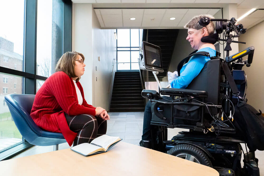 an adult in a wheelchair with a mounted AAC device interacting with a young adult near a stairwell