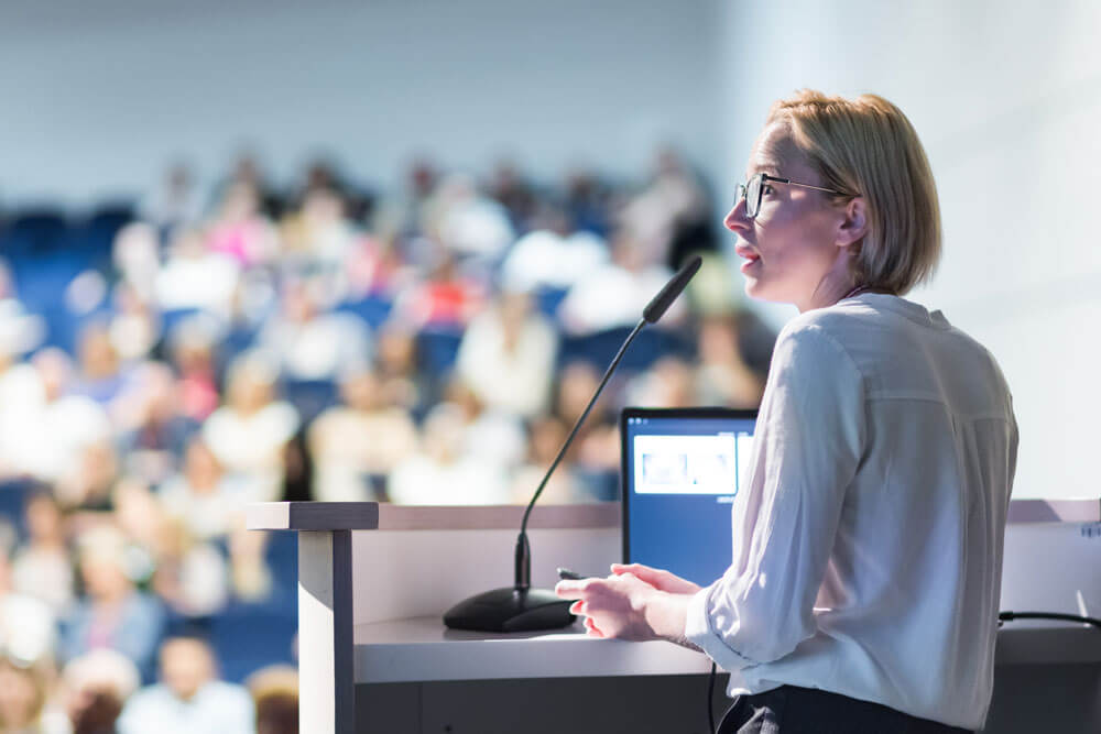 a person standing at a lectern in front of a conference room full of people