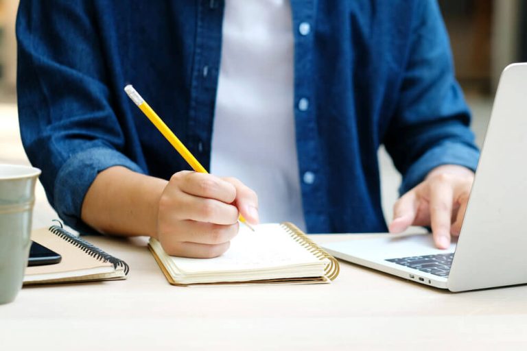 close up of a person sitting in front of a laptop writing in a notebook